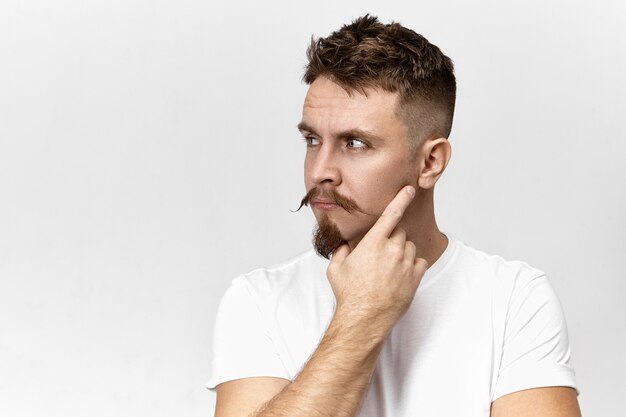 Jovem barbudo elegante e atraente com bigode pensando em algo, segurando a mão no rosto, posando contra o fundo da parede do estúdio em branco com espaço de cópia para suas informações de publicidade