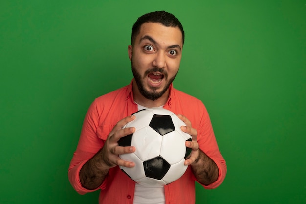 Foto grátis jovem barbudo emocional com camisa laranja segurando uma bola de futebol e olhando gritando em pé sobre a parede verde