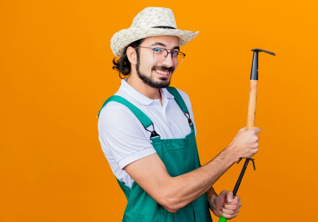 Jovem barbudo jardineiro usando macacão e chapéu segurando enxadão e mini ancinho olhando para a frente sorrindo em pé sobre a parede laranja