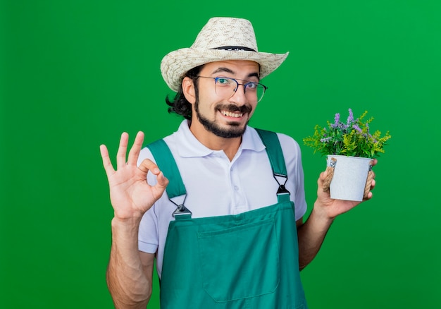 Jovem barbudo jardineiro usando macacão e chapéu segurando um vaso de plantas sorrindo e mostrando sinal de ok