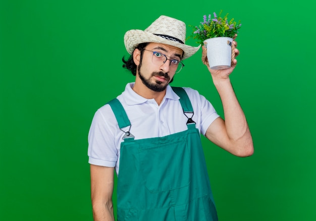 Jovem barbudo jardineiro, vestindo macacão e chapéu, segurando um vaso de plantas perto da cabeça, perplexo