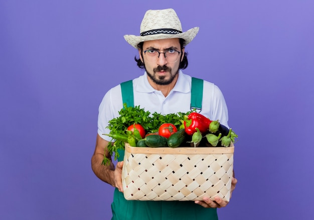 Jovem barbudo jardineiro, vestindo macacão e chapéu, segurando uma caixa cheia de vegetais, olhando para a frente com uma cara séria e carrancuda em pé sobre a parede azul