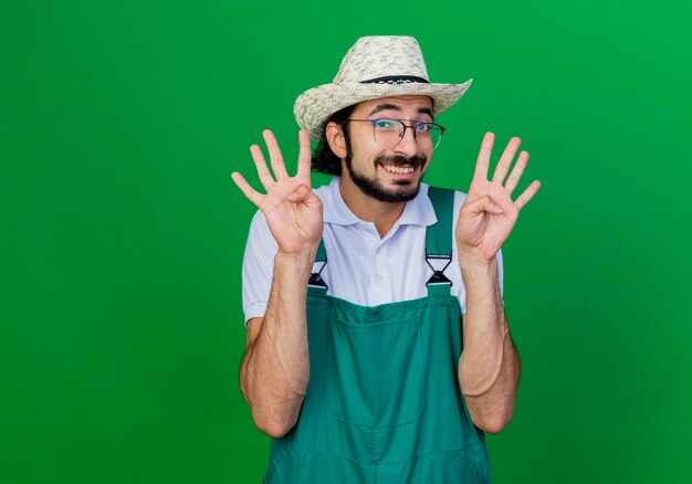 Jovem barbudo jardineiro vestindo macacão e chapéu sorrindo e mostrando o número oito