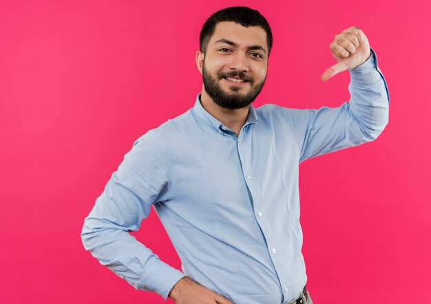 Jovem barbudo satisfeito com uma camisa azul apontando para si mesmo, sorrindo confiante