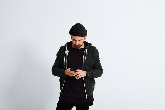 Jovem barbudo vestido com roupas casuais pretas e marinho olha para seu smartphone isolado no branco