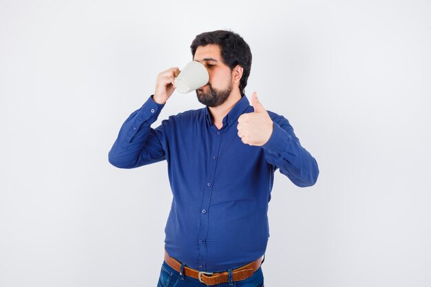 Jovem, bebendo um copo d'água e aparecendo o polegar em uma camisa azul e calça jeans e olhando otimista. vista frontal.