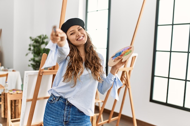 Foto grátis jovem bela artista hispânica sorrindo confiante segurando pincel e paleta no estúdio de arte