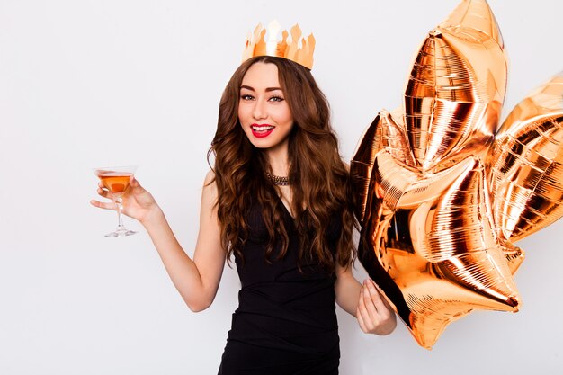 Jovem bela mulher comemorando no vestido preto sorrir e posando com cocktail na mão e balões de pureza.