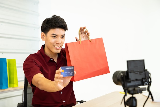 Jovem blogueiro comerciante on-line masculino asiático usando câmera para mostrar vídeo ao vivo sacola de compras e cartão de crédito Mídia social Influenciador de compras on-line e conceito de pagamento