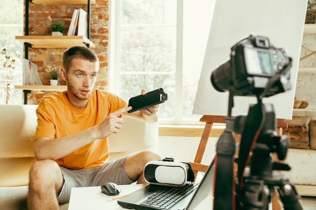 Jovem blogueiro do sexo masculino, caucasiano, com equipamento profissional, gravação de revisão de vídeo de óculos vr em casa blogging, videoblog, vlogging. o homem avalia um fone de ouvido de realidade virtual durante a transmissão ao vivo.