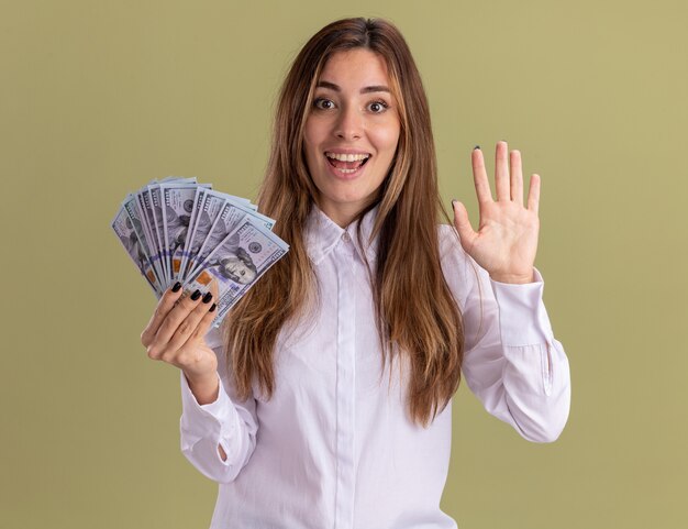 Jovem bonita caucasiana animada segurando dinheiro e gesticulando cinco em verde oliva