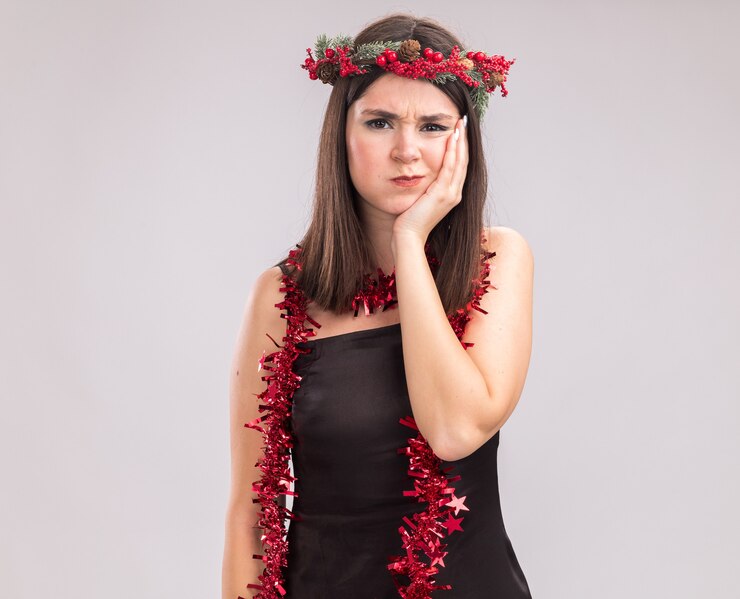 Foto grátis jovem, bonita, caucasiana, carrancuda, usando coroa de flores de natal e guirlanda de ouropel em volta do pescoço, olhando para a câmera, mantendo as mãos no rosto com bochechas inchadas