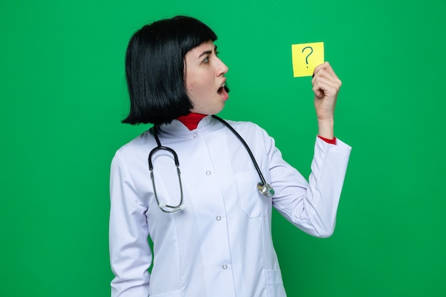 Jovem bonita caucasiana chocada com uniforme de médico e estetoscópio segurando e olhando para o bilhete interrogativo