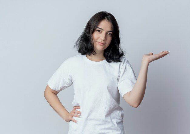 Jovem bonita caucasiana confiante colocando a mão na cintura e mostrando a mão vazia isolada no fundo branco