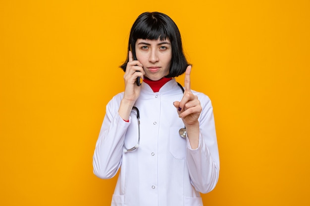 Jovem bonita caucasiana confiante com uniforme de médico com estetoscópio falando no telefone e apontando para cima