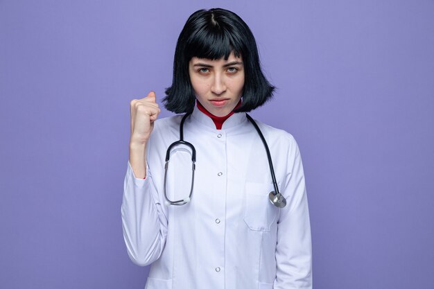 Jovem bonita caucasiana confiante em uniforme de médico com estetoscópio, segurando o punho e olhando para frente