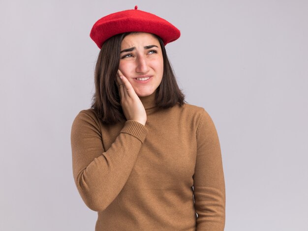 Jovem bonita caucasiana dolorida com chapéu boina coloca a mão no rosto e olha para o lado em branco