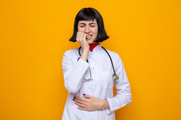 Jovem, bonita, caucasiana, dolorida, com uniforme de médico e estetoscópio, colocando a mão na barriga e roendo as unhas