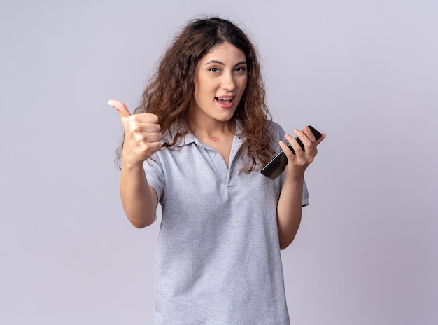 Jovem, bonita, caucasiana, impressionada, segurando um celular e mostrando o polegar