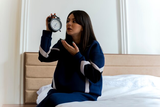 Jovem, bonita, caucasiana, impressionada, sentada na cama no quarto segurando e olhando para o despertador e apontando com a mão para ele