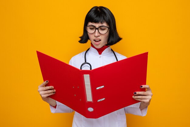 Jovem, bonita, caucasiana, insatisfeita, com óculos, uniforme de médico, estetoscópio segurando e olhando para a pasta de arquivo