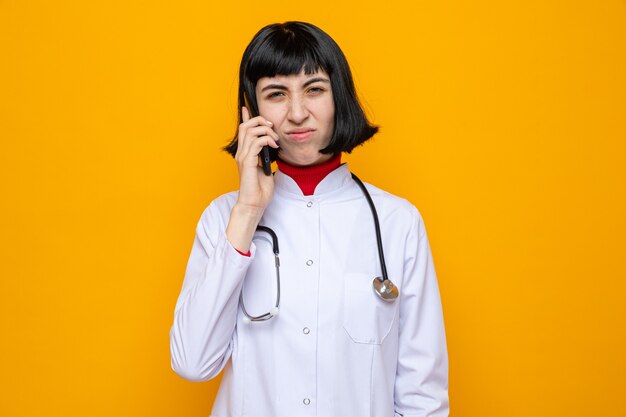 Jovem bonita caucasiana insatisfeita com uniforme de médico e estetoscópio falando ao telefone