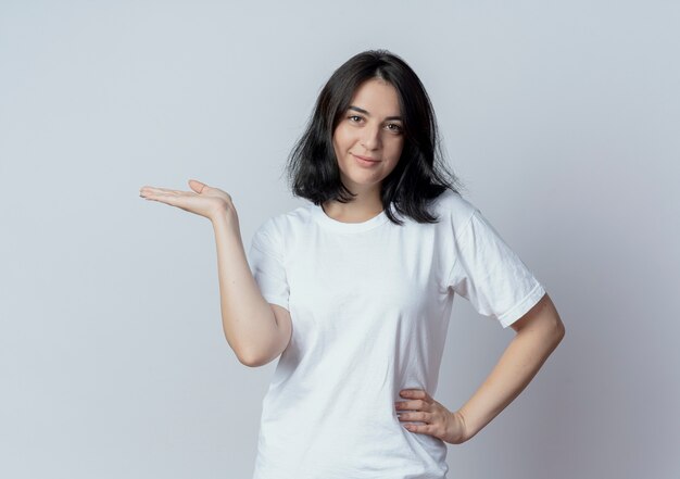 Jovem, bonita, caucasiana, satisfeita, colocando a mão na cintura e mostrando a mão vazia, isolada no fundo branco