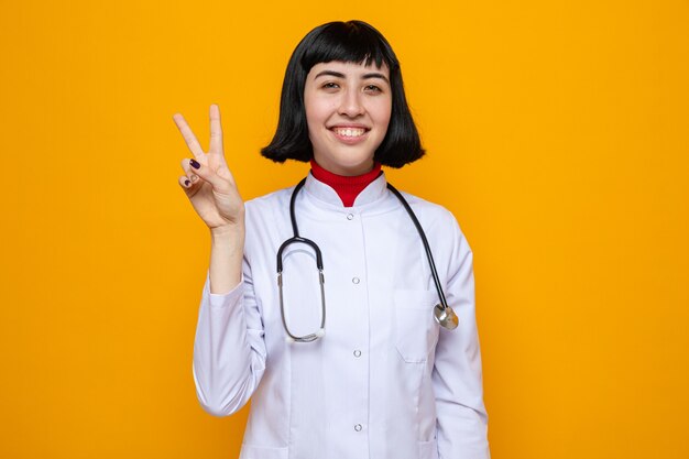 Jovem, bonita, caucasiana, satisfeita, com uniforme de médico e estetoscópio gesticulando sinal de vitória