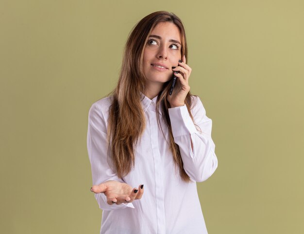 Jovem, bonita, caucasiana, satisfeita, mantém a mão aberta e fala ao telefone, olhando para o lado isolado na parede verde oliva com espaço de cópia