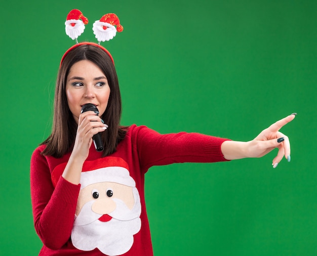Jovem, bonita, caucasiana, satisfeita, usando blusa de Papai Noel e bandana, falando ao microfone, olhando e apontando para o lado isolado na parede verde