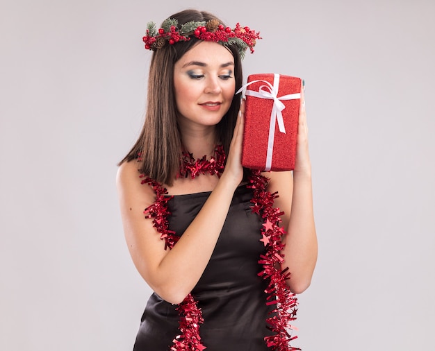 Jovem, bonita, caucasiana, satisfeita, usando coroa de flores de natal e guirlanda de ouropel em volta do pescoço segurando e olhando para um pacote de presente