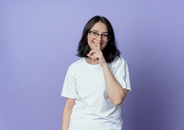 Jovem, bonita, caucasiana, sorridente, usando óculos, gesticulando para o silêncio