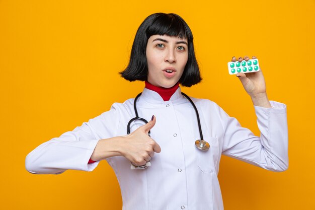 Jovem bonita caucasiana surpresa com uniforme de médico com estetoscópio segurando a embalagem do comprimido e apontando para cima