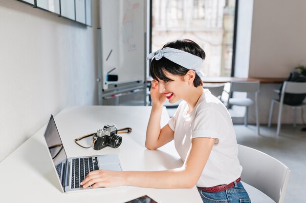 Jovem bonita com cabelo escuro em roupa retrô, trabalhando no laptop em casa, sentada à mesa