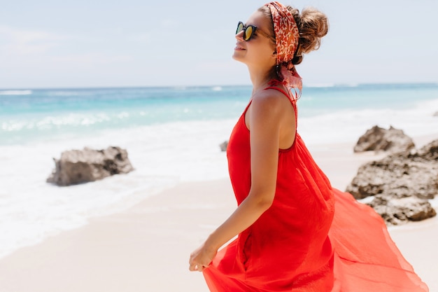 Jovem bonita com pele levemente bronzeada, posando com um sorriso na costa do mar. Modelo feminino fascinante, relaxando na natureza em um dia ensolarado.