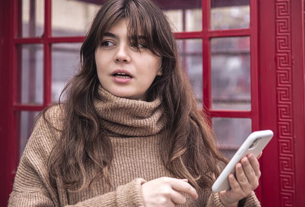 Jovem bonita com um smartphone no fundo de uma cabine telefônica vermelha.