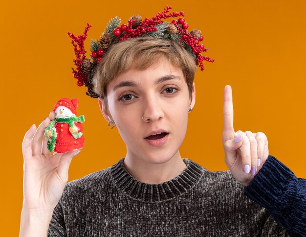 Jovem bonita com uma coroa de flores de natal e segurando uma estátua de boneco de neve de natal olhando para a câmera apontando para cima isolada em fundo laranja