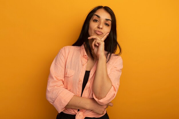 Jovem bonita confiante vestindo camiseta rosa colocando a mão no queixo isolado em amarelo com espaço de cópia