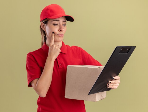 Foto grátis jovem bonita confusa entregadora de uniforme segurando uma caixa de papelão e olhando para a prancheta isolada na parede verde oliva