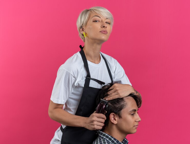 Foto grátis jovem bonita e confiante barbeiro feminino de uniforme segurando ferramentas de barbeiro e cortando o cabelo de menino isolado na parede rosa