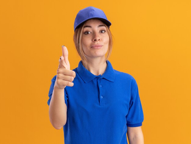 Jovem bonita e confiante entregadora de uniforme aponta para a câmera em laranja