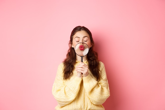 Jovem bonita e romântica amplia seus lábios com lupa, fazendo careta de beijo, em pé com os olhos fechados contra um fundo rosa