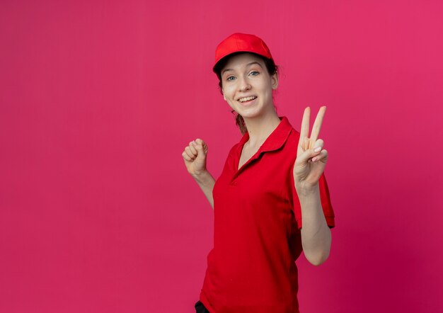 Jovem bonita entregadora alegre de uniforme vermelho e boné em pé na vista de perfil, cerrando os punhos e fazendo o sinal da paz, isolado no fundo carmesim com espaço de cópia