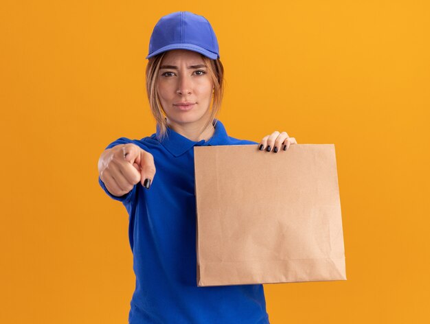 Jovem bonita entregadora de uniforme confiante segurando um pacote de papel e pontos na frente isolados na parede laranja