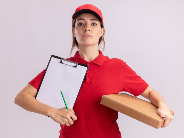 Jovem bonita entregadora de uniforme confiante segurando uma caixa de pizza e uma prancheta isoladas na parede branca