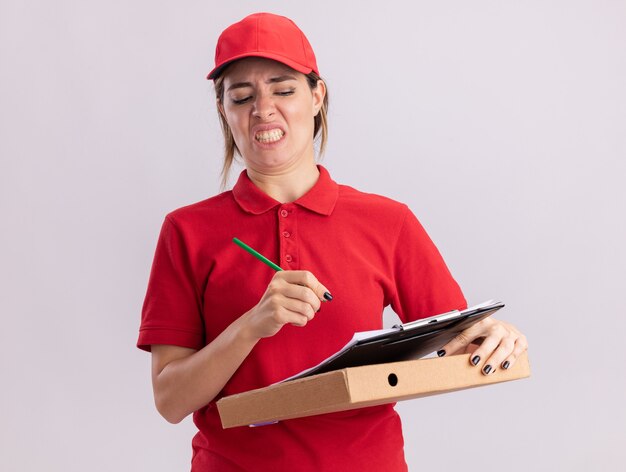 Jovem bonita entregadora de uniforme descontente segurando lápis e prancheta na caixa de pizza isolada na parede branca