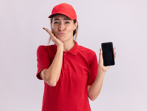 Jovem bonita entregadora de uniforme empolgada coloca a mão no queixo e segura o telefone isolado na parede branca