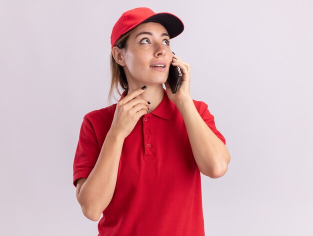 Jovem bonita entregadora de uniforme impressionada fala ao telefone e olha para o lado isolado na parede branca