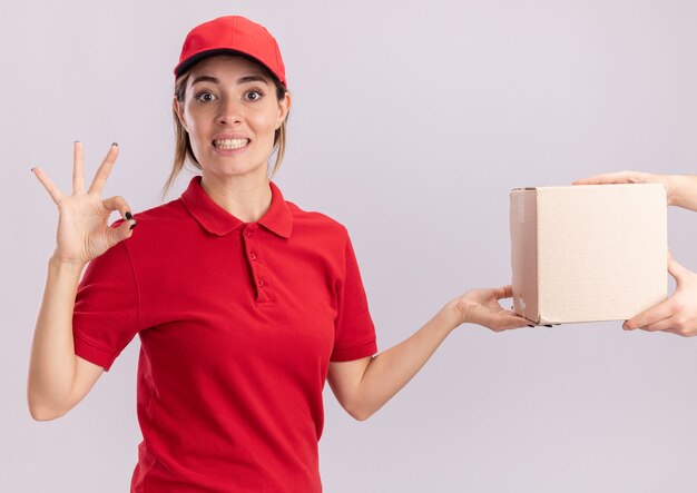 Jovem, bonita, entregadora de uniforme, impressionada, gesticula com a mão ok e dá uma caixa de papelão para alguém branco