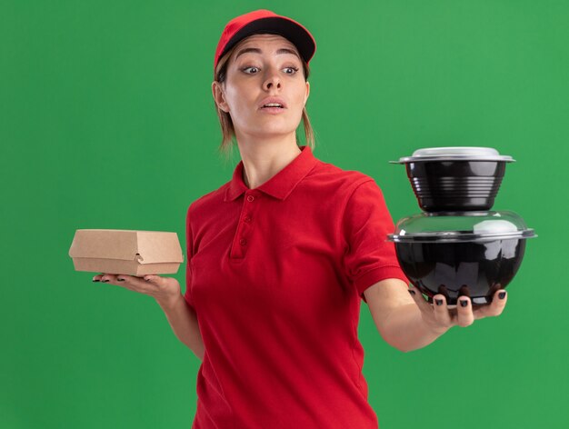 Jovem, bonita, entregadora de uniforme, impressionada segurando um pacote de comida e olhando para os recipientes de comida no verde